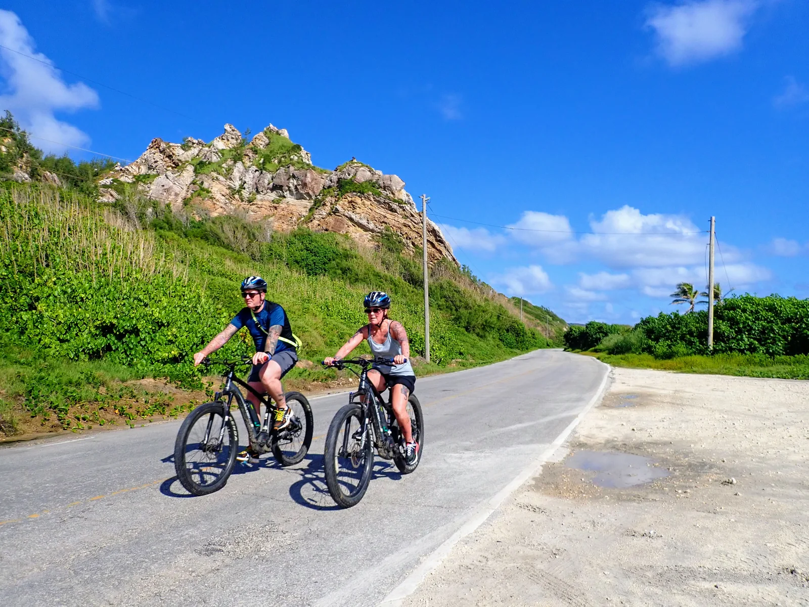 Two cyclists on an eBike Barbados ride along a countryside road beside a rocky hillside